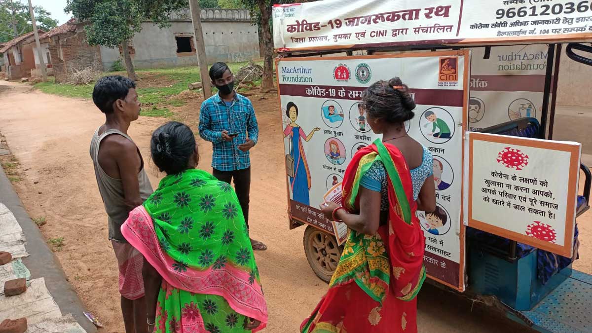 People gathered around a mobile cart displaying COVID-19 awareness posters in a rural setting.