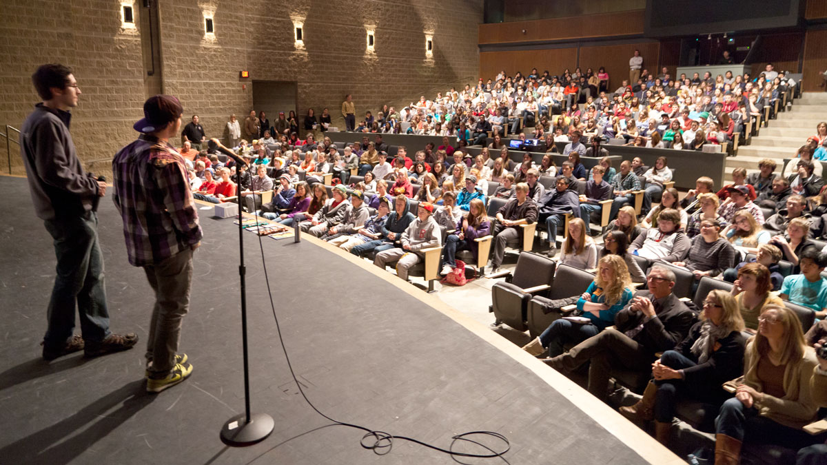 A crowded auditorium with two speakers on stage addressing the audience.