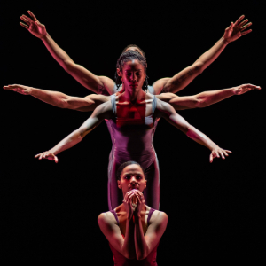 Four dancers aligned in a line, creating an optical illusion of multiple arms, on a dark stage with dramatic lighting.