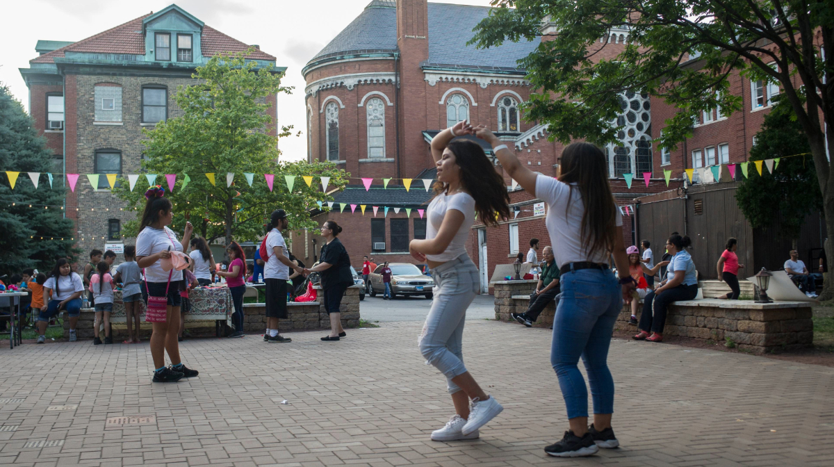 A diverse group of people talking, eating, and dancing at a celebration in a church courtyard.