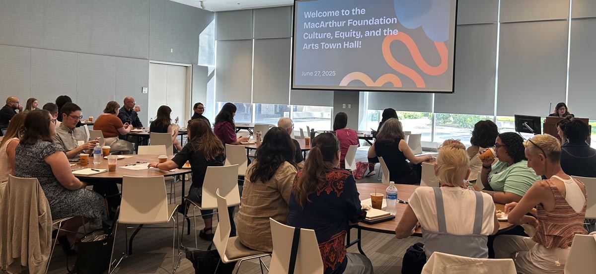 Attendees seated at tables in a conference room with a large screen displaying a welcome message for the MacArthur Foundation Culture, Equity, and the Arts Town Hall.