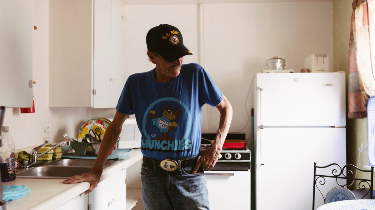 Man in a blue T-shirt and U.S. Navy cap stands in a kitchen next to a stove and refrigerator, with hands on the counter.