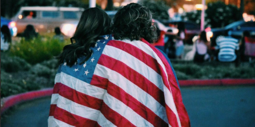 Two people wrapped in a large American flag stand together outdoors with a crowd in the background.