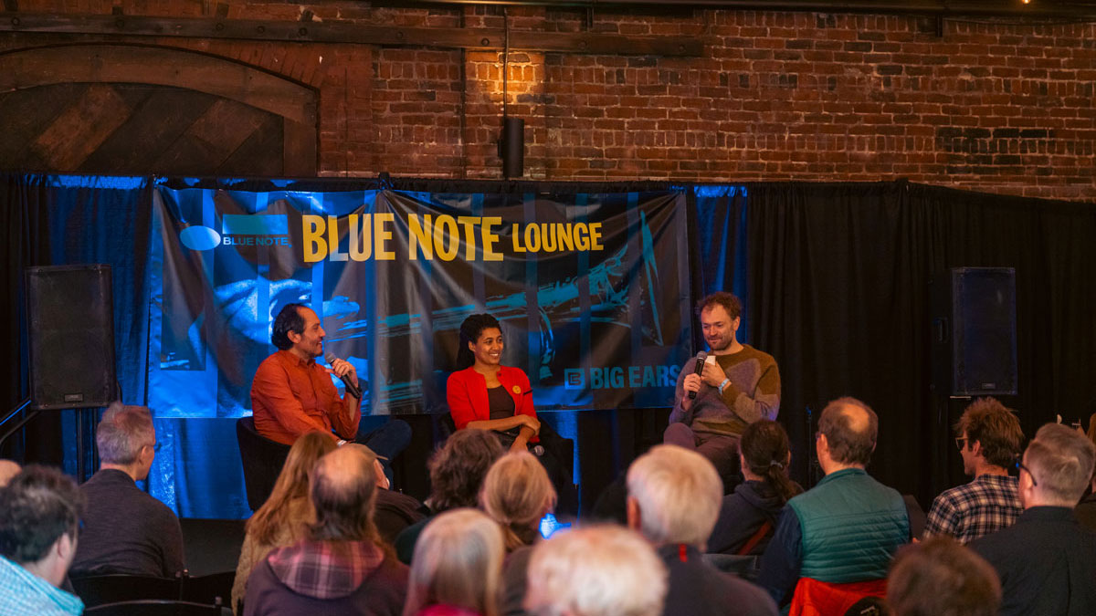 Three people sit in a row and answer questions from an audience in front of them. There is a sign behind them that reads “Blue Note Lounge.” 