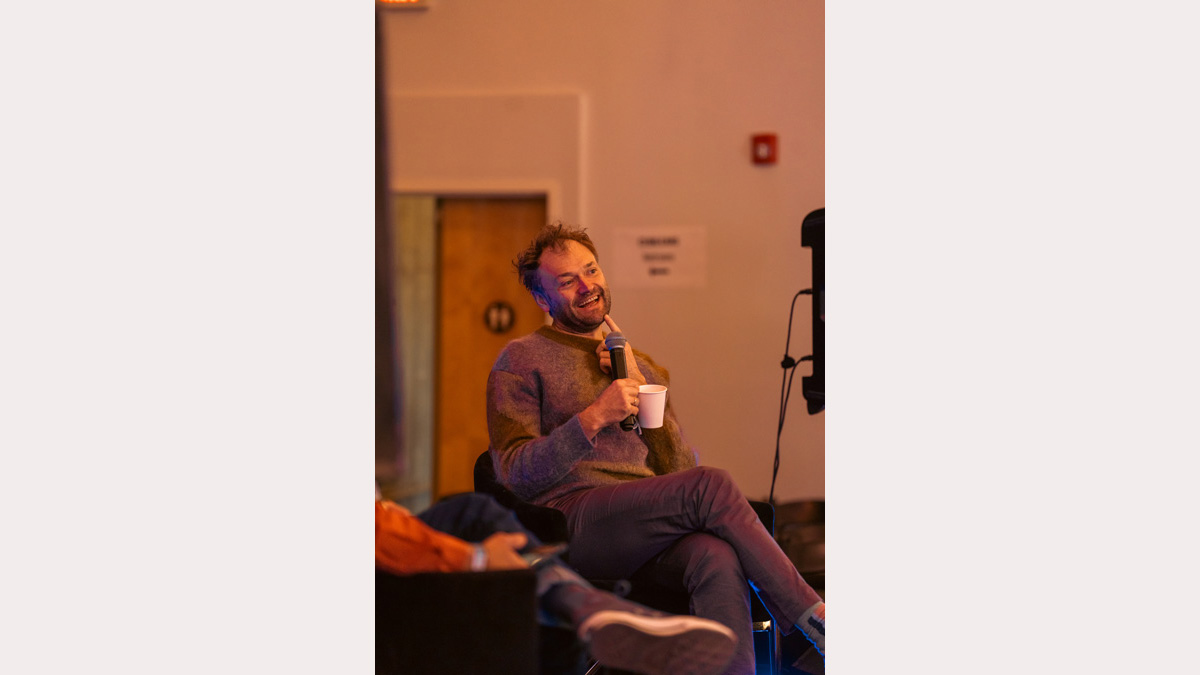 A close photo of a bearded man with a playful expression. He sits during an interview at a music festival. 