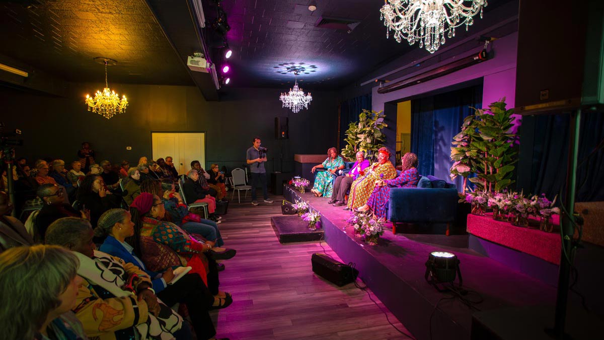 Four women sit on a stage, deep in conversation. A crowd sits in front of them, listening attentively to the conversation.