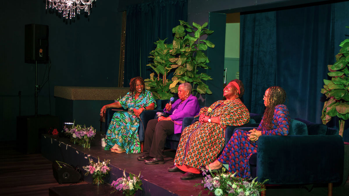 Close on a panel of four women, seated on a small stage. The Women sit in blush velvet chairs surrounded by bouquets of flowers.