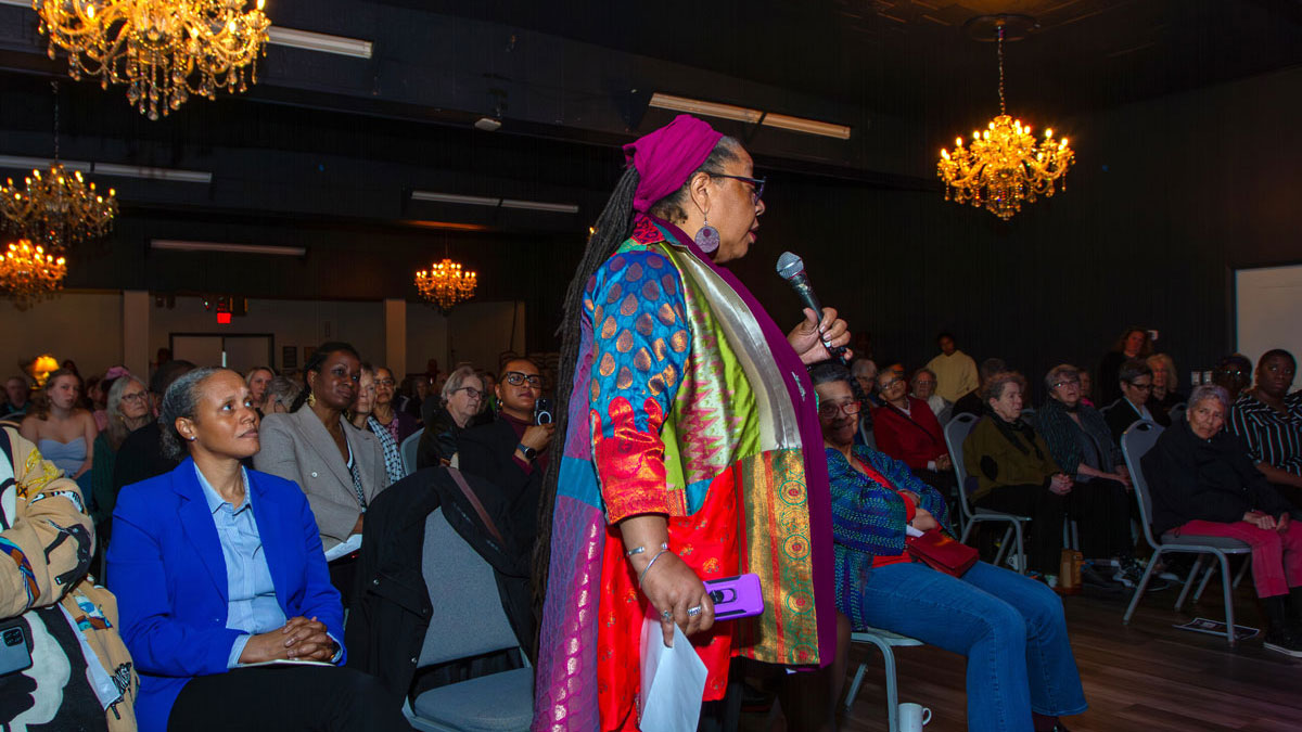A woman stands, holding a microphone, to ask a question at a public event. Behind her, and attentive crowd sits listening.