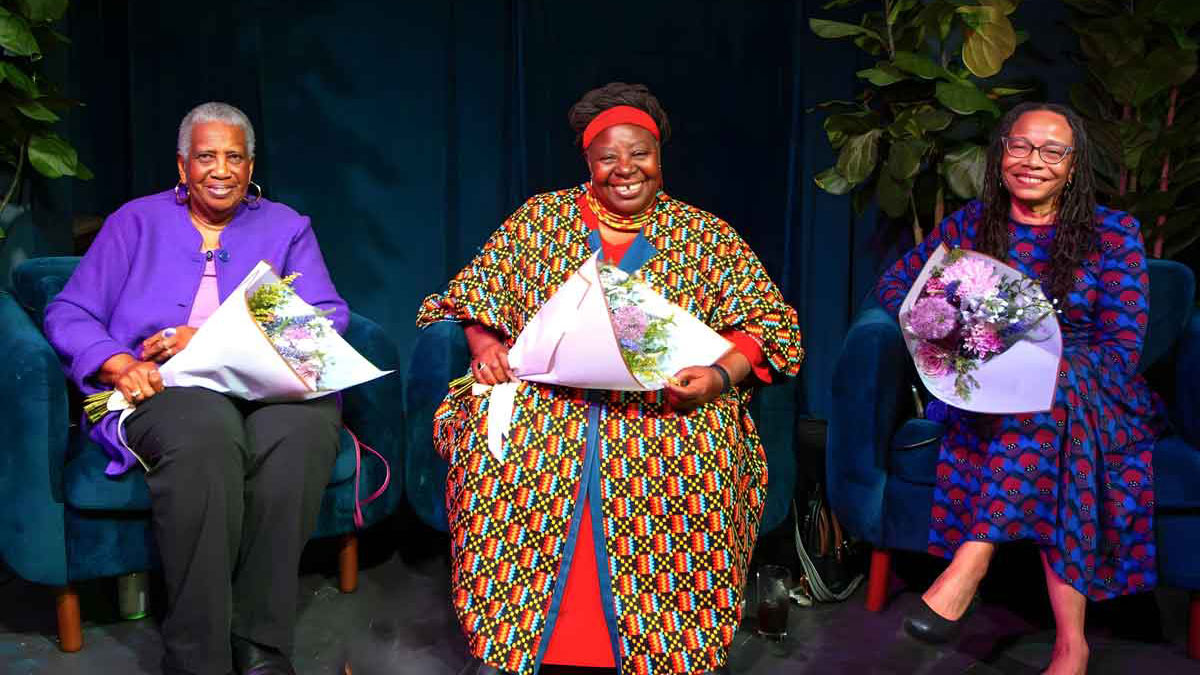 A trio of smiling women sit holding bouquets of light purple flowers, after being honored at an event. 