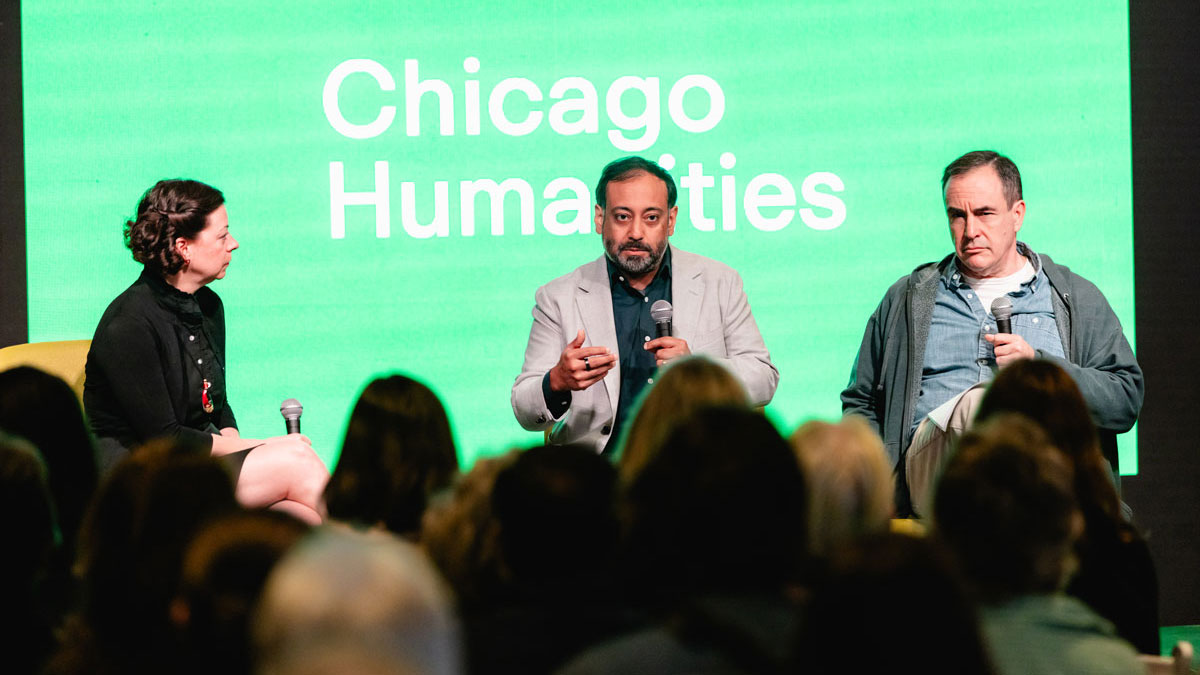 A woman interviews two men in front of an attentive audience. All three hold microphones and sit in front of a bright green screen that reads “Chicago Humanities.”