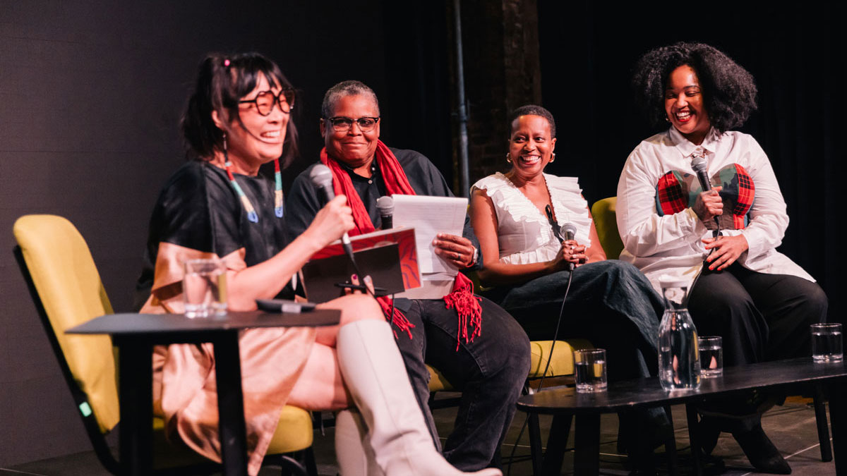 A quartet of women sit in a row of chairs for a panel discussion. Each woman holds a microphone. All four are laughing, mid-discussion. 