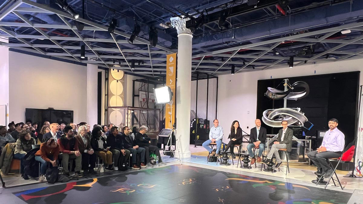 A crowd of people sits in a museum atrium, watching a demonstration by five prominent mathematicians. 
