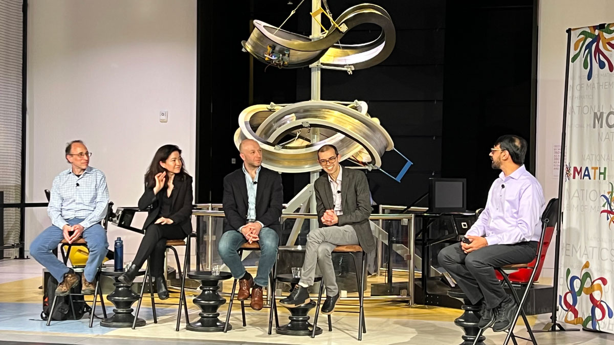Five mathematicians sit in a row during a panel discussion at a museum. Behind them is a large poster displaying a complex geometric shape. 