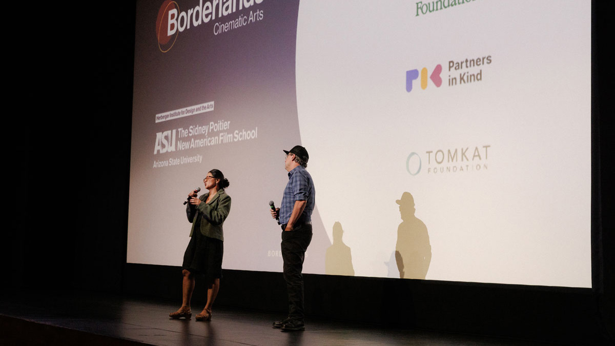 A man and woman stand holding microphones to introduce a film screening. Behind them is a large movie screen, displaying the sponsors of the film.