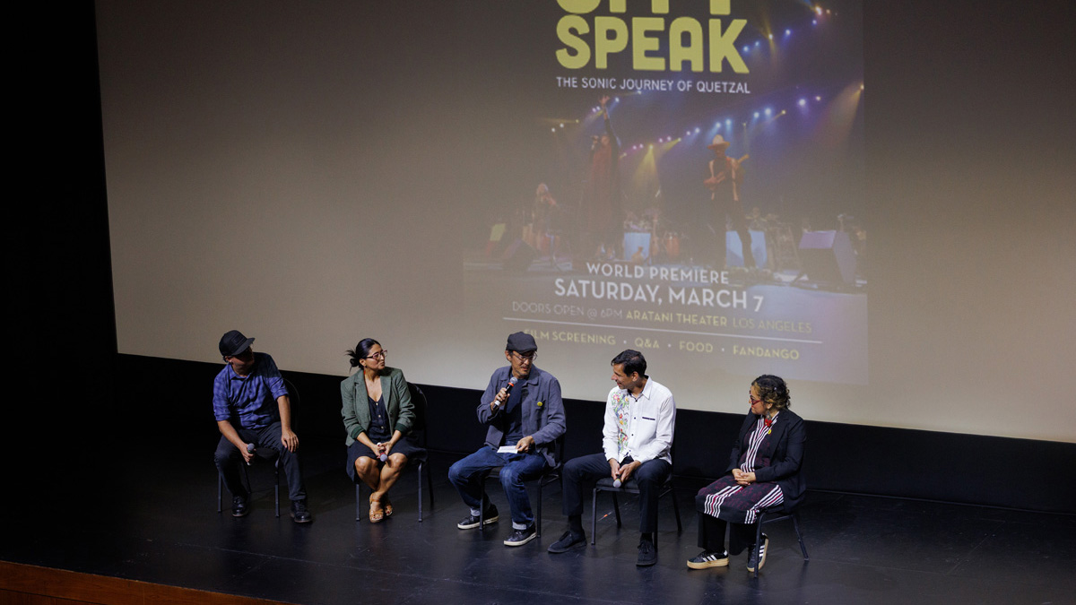 Five men and women sit in a row, holding microphones. Behind them is a movie screen displaying the poster for the documentary Let the City Speak: The Sonic Journey of Quetzal. 