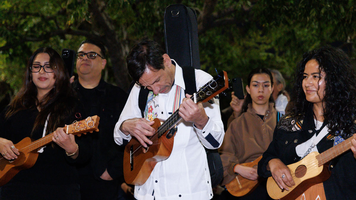 A man in a white button-down strums a vihuela while surrounded by men and women with similar instruments. The man’s head is down, focusing on the music. 