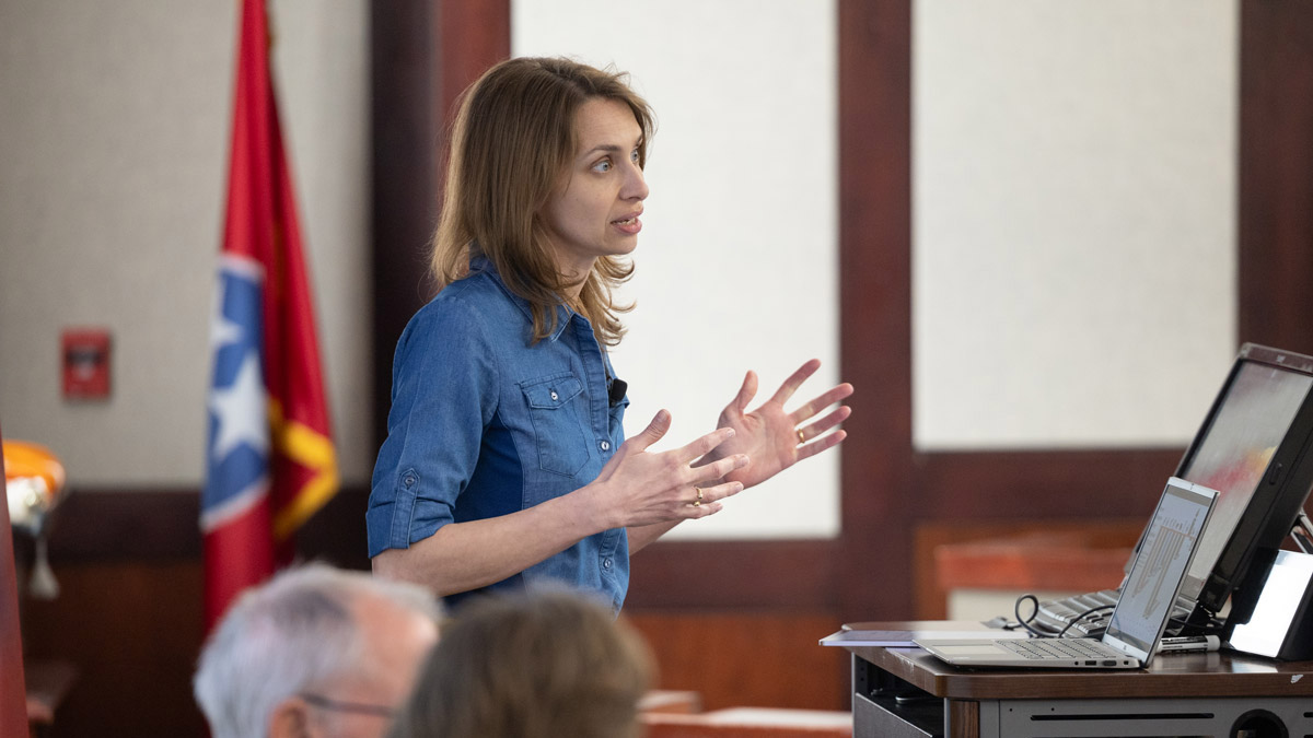 A photograph of a woman from the waist up as she stands behind a podium, mid-lecture. The woman gestures emphatically with her hands to make a point.