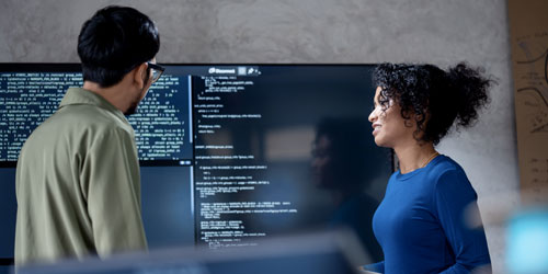 Two people discuss in front of a screen displaying computer code.