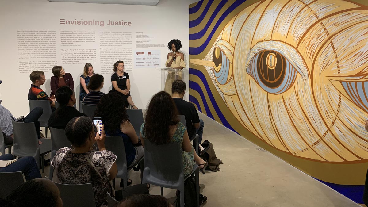 A speaker stands at a podium beside a mural with a stylized eye; audience and panelists listen in a gallery.