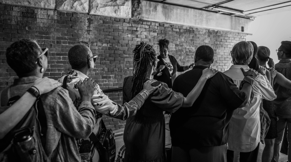 A group of people stand with backs to the camera and arms across each other's shoulders.