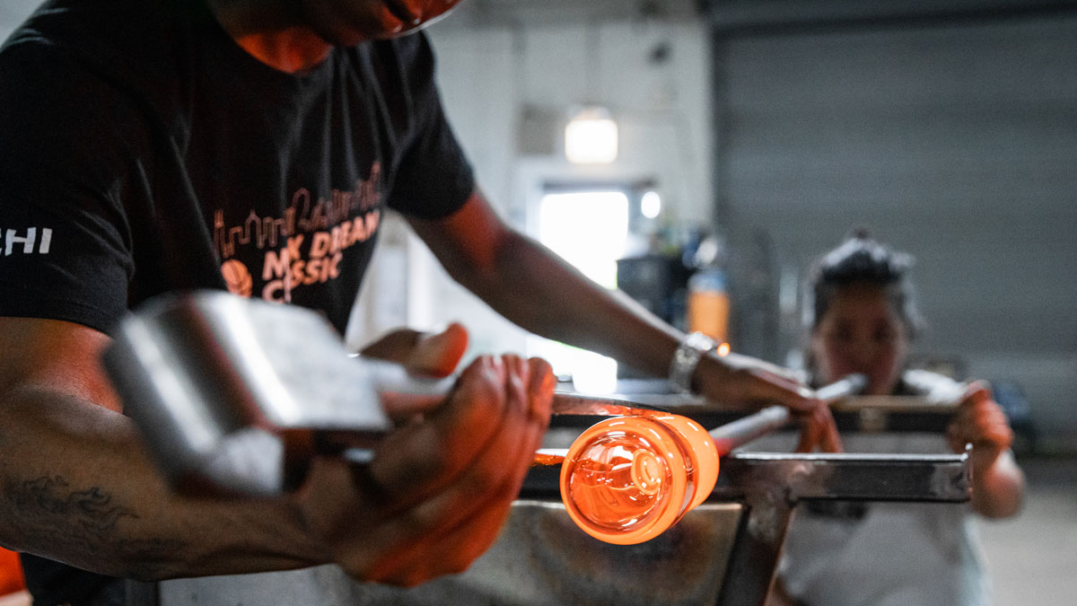 Person glassblowing with glowing orange glass orb and assisting individual in a workshop.