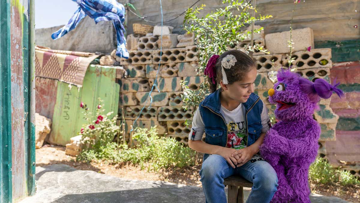 A young girl sits outside on a stool, smiling at a purple puppet. They are surrounded by cinder blocks and some plants.
