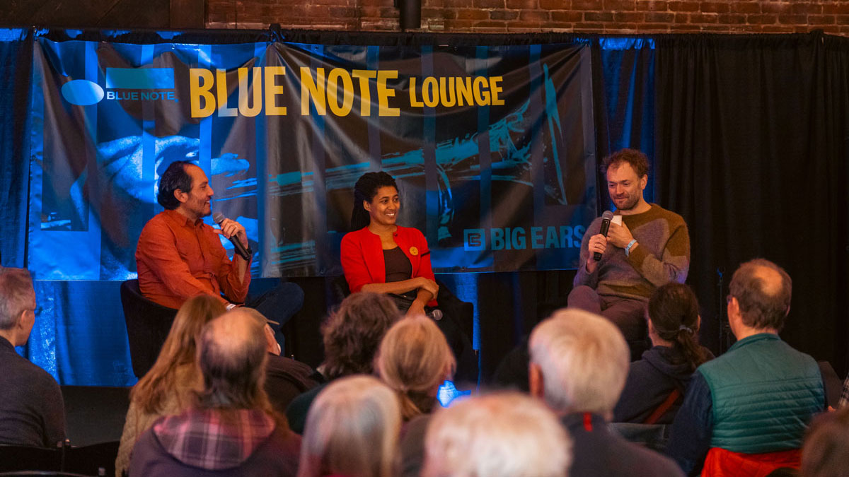 Three people sit in a row and answer questions from an audience in front of them. There is a sign behind them that reads “Blue Note Lounge.” 
