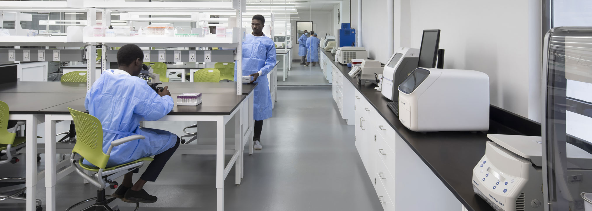 A modern laboratory with two individuals in blue lab coats working at benches with scientific equipment.