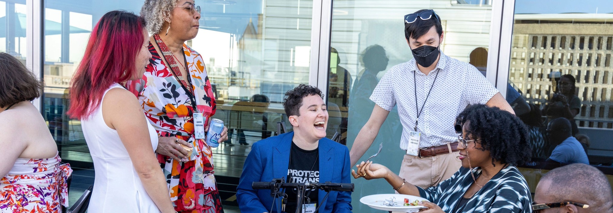 A diverse group of people socialize and laugh outdoors; one person sits in a wheelchair, another wears a mask.