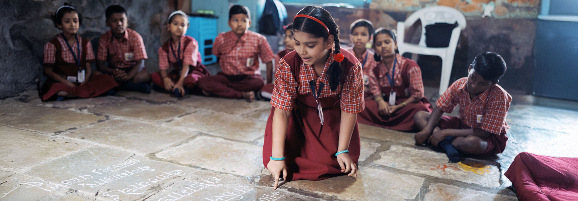 A girl writes on the classroom floor with chalk while surrounded by seated classmates in red plaid uniforms.