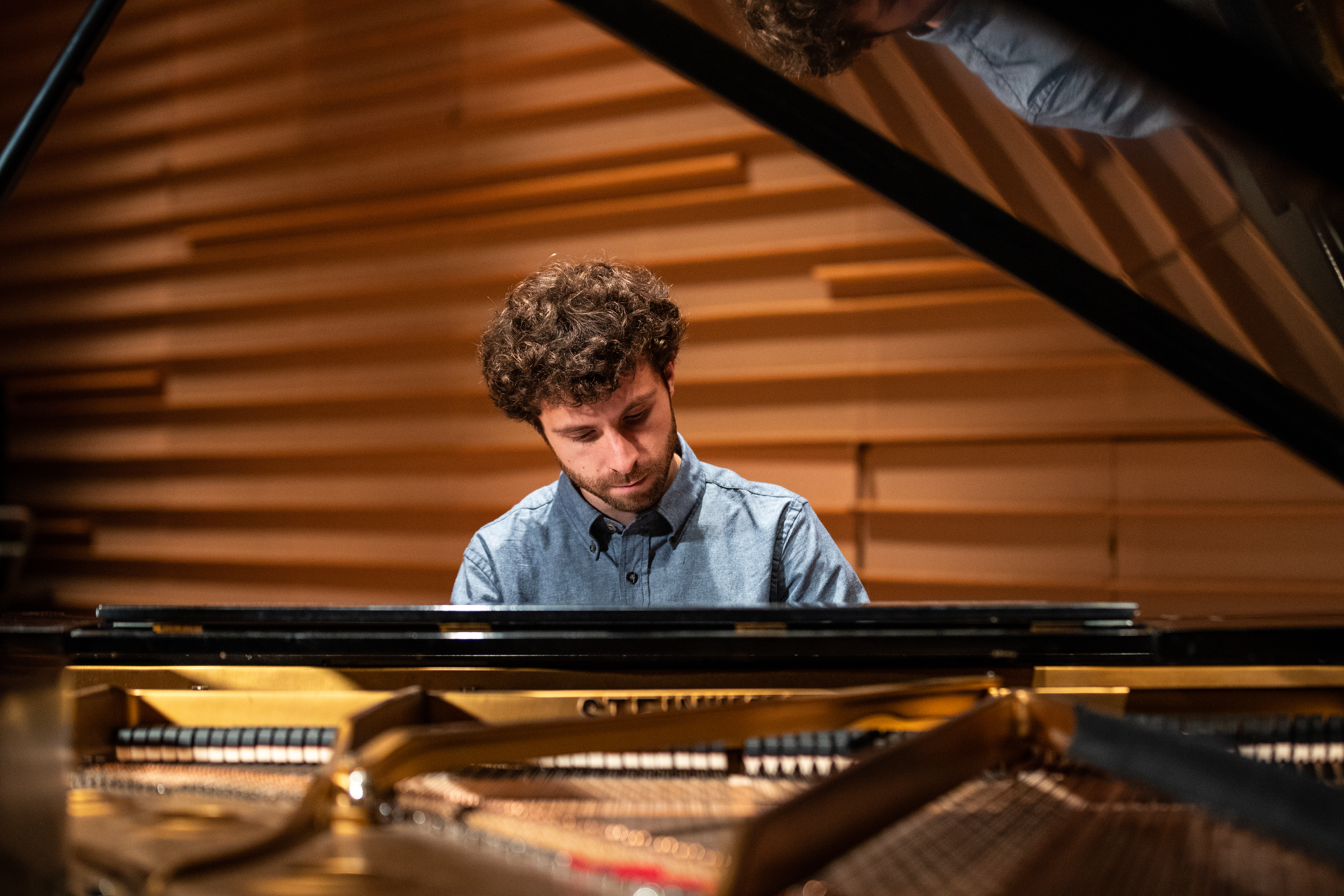 A man with brown hair in a blue and white shirt plays the piano.