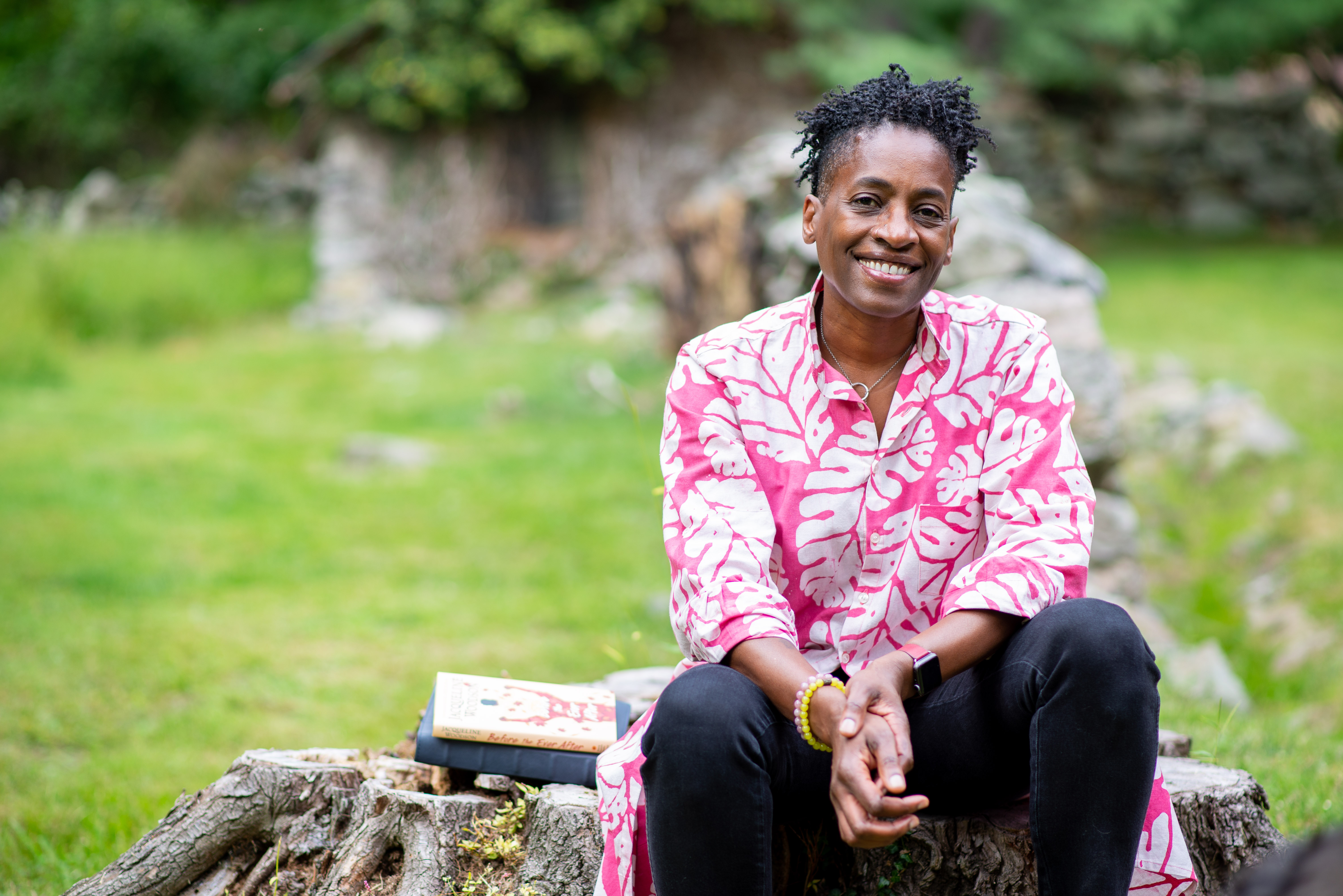 A woman sits on a tree stump with books beside her.