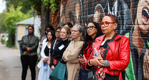 A group of people stand attentively in front of a mural on a brick wall outdoors.
